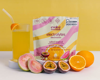 A package of evolve essentials electrolytes in 'salted tropical' flavor, styled with passionfruit, sliced guavas, and oranges on a white surface against a yellow background.